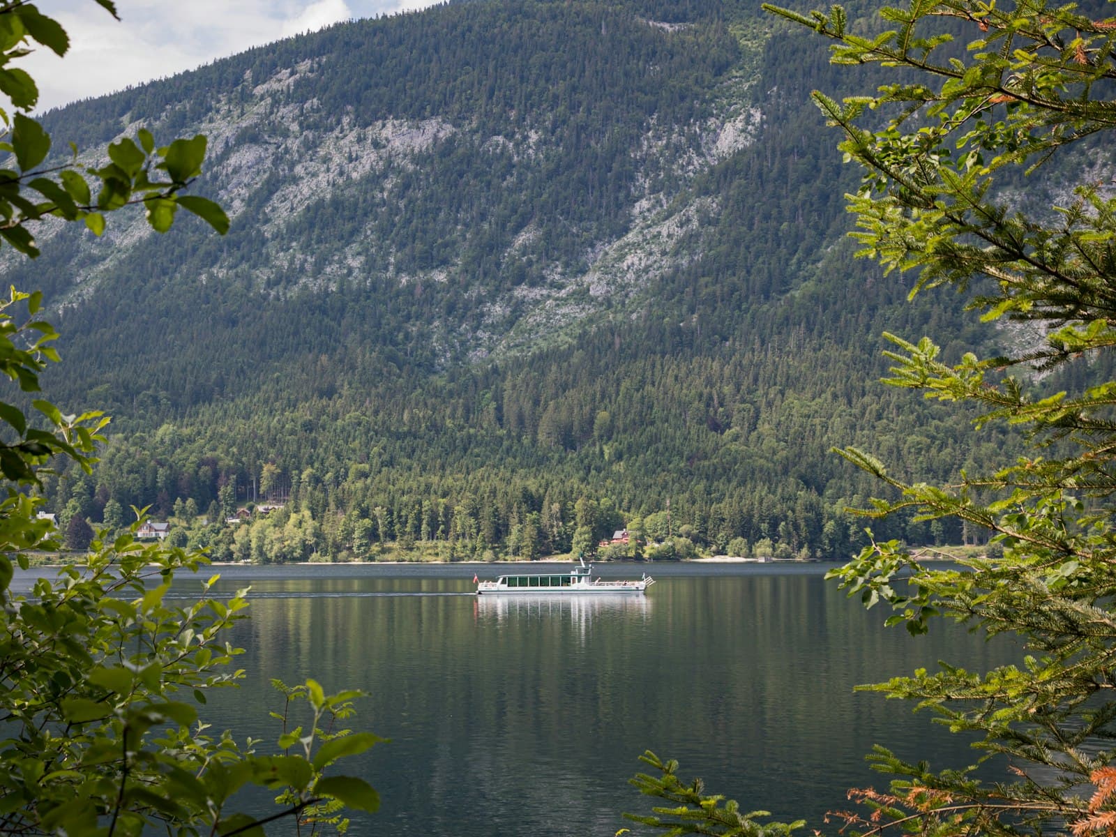 A white passenger boat crossing Lake Altaussee below the wooded slopes of the Loser, seen through branches on the shore.