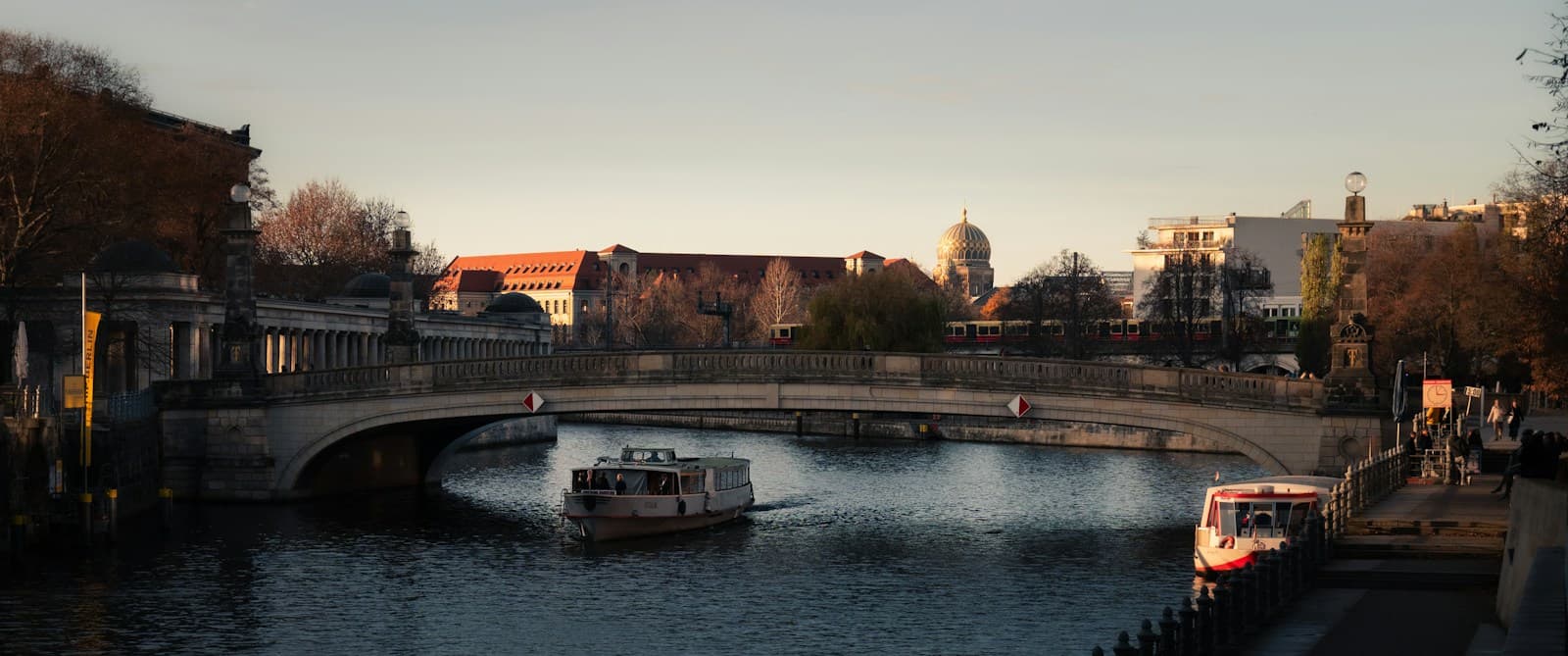 Ein Ausflugsschiff fährt auf der Spree unter der Friedrichsbrücke durch. Die Bäume am Ufer sind schon herbstlich, hinten glänzt die Kuppel der Neuen Synagoge.