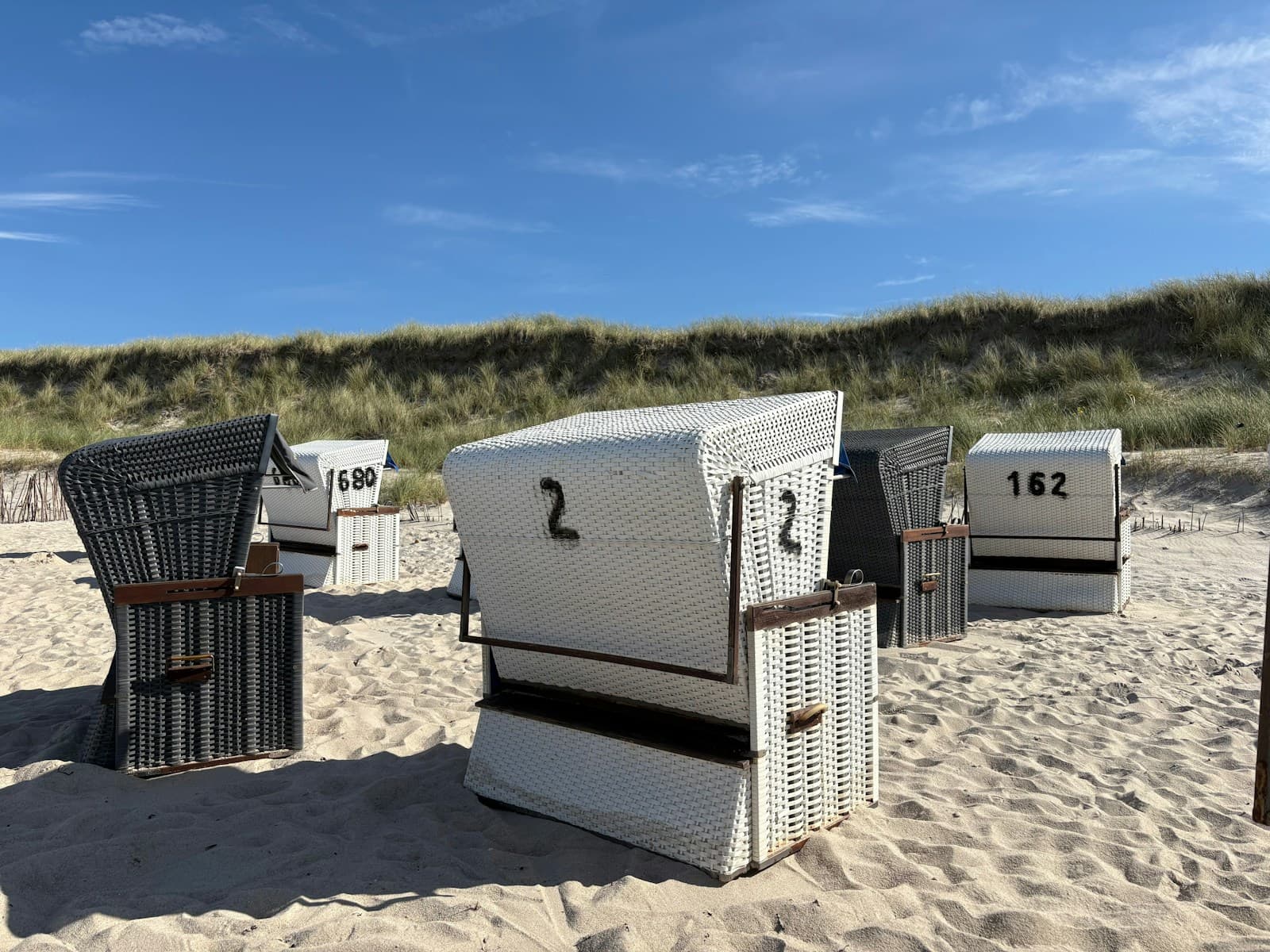 Numbered wicker beach chairs with their hoods closed, standing in pale sand on Sylt, a dune of long grass behind them and a clear blue sky above.